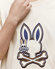 Someone wearing the Psycho Bunny KIDS WADE GRAPHIC TEE—KT0200054, a cream-colored 100% cotton shirt with a large embroidered Bunny head above crossed bones and a smaller bunny skull and crossbones on the left side.