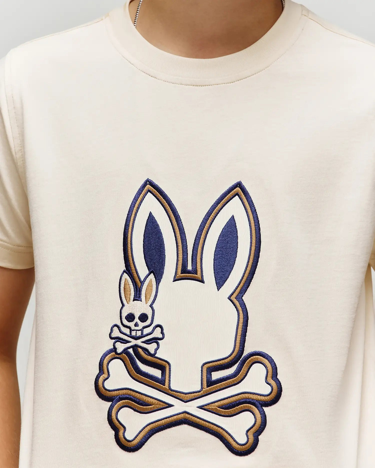 A person wearing the Psycho Bunny KIDS WADE GRAPHIC TEE (KT0200054), a cream 100% cotton shirt with a large bunny head above crossed bones graphic and a small embroidered Bunny with skull and crossbones.
