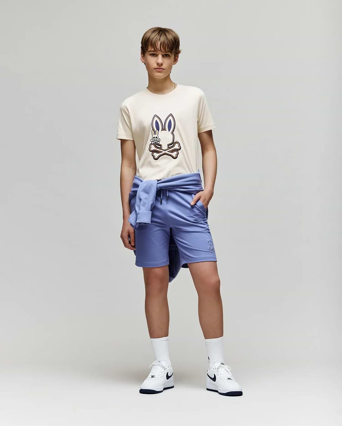 A teenage boy stands against a plain background wearing the Psycho Bunny KIDS WADE GRAPHIC TEE - KT0200054 in beige with an embroidered bunny, paired with light blue shorts, a matching sweatshirt tied around his waist, white socks, and Nike sneakers.