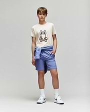 A teenage boy stands against a plain background wearing the Psycho Bunny KIDS WADE GRAPHIC TEE - KT0200054 in beige with an embroidered bunny, paired with light blue shorts, a matching sweatshirt tied around his waist, white socks, and Nike sneakers.