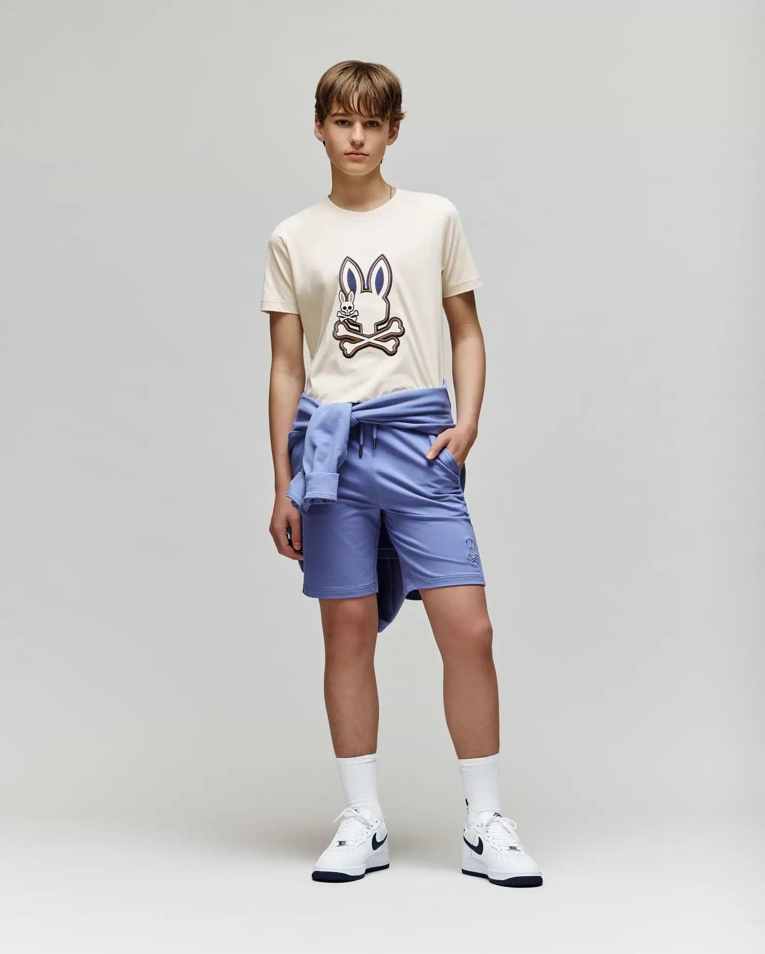 A teenage boy stands against a plain background wearing the Psycho Bunny KIDS WADE GRAPHIC TEE - KT0200054 in beige with an embroidered bunny, paired with light blue shorts, a matching sweatshirt tied around his waist, white socks, and Nike sneakers.