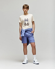 A teenage boy stands against a plain background wearing the Psycho Bunny KIDS WADE GRAPHIC TEE - KT0200054 in beige with an embroidered bunny, paired with light blue shorts, a matching sweatshirt tied around his waist, white socks, and Nike sneakers.