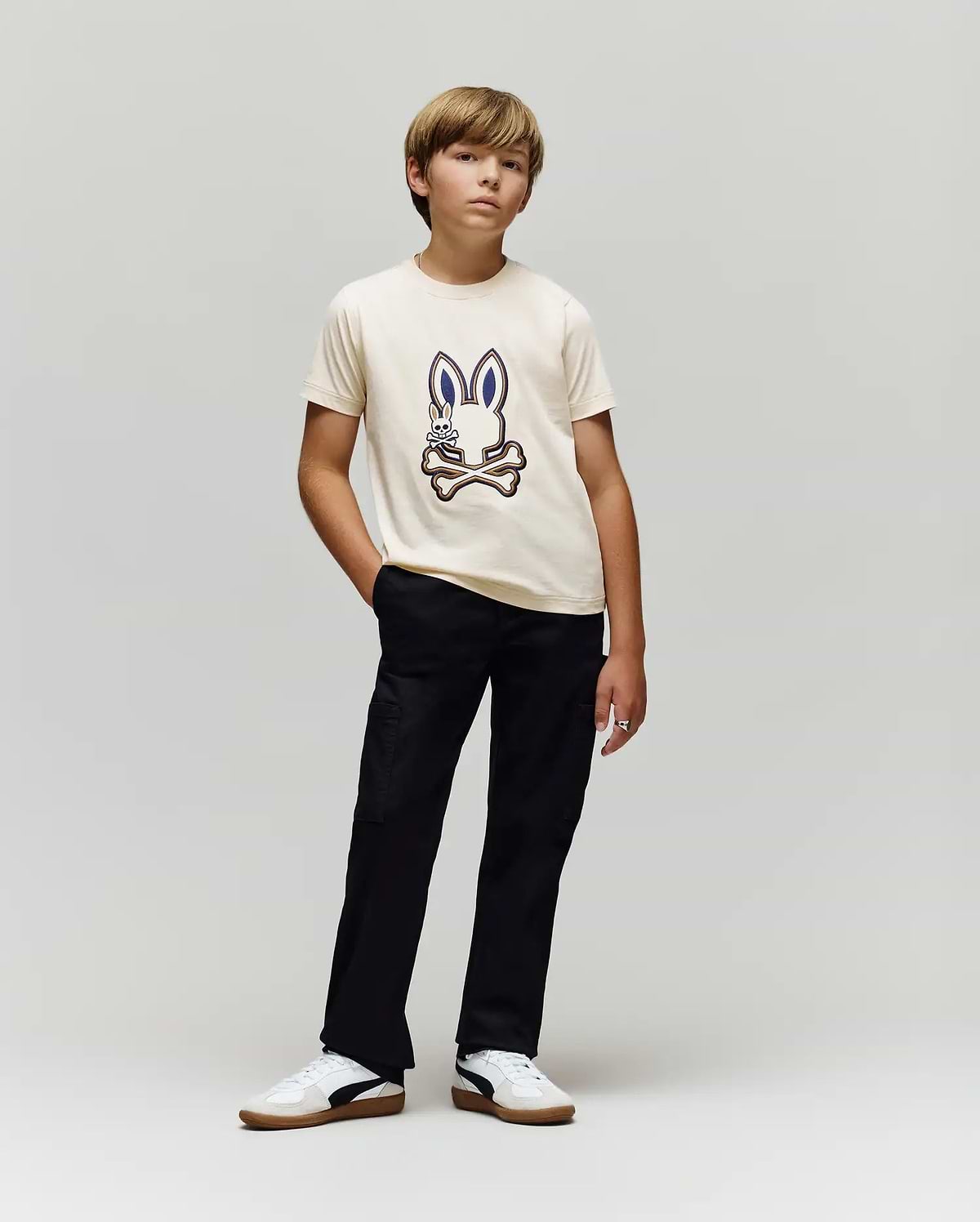 A boy poses against a plain background, wearing the Psycho Bunny KIDS WADE GRAPHIC TEE - KT0200054 in cream, made from 100% cotton with an embroidered Bunny and crossed bones, paired with black pants and white sneakers.