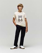 A boy poses against a plain background, wearing the Psycho Bunny KIDS WADE GRAPHIC TEE - KT0200054 in cream, made from 100% cotton with an embroidered Bunny and crossed bones, paired with black pants and white sneakers.