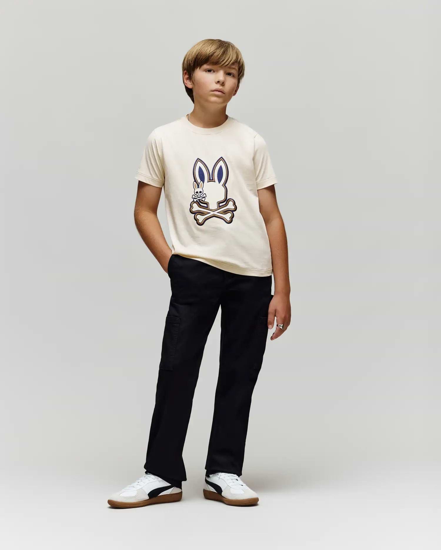 A boy poses against a plain background, wearing the Psycho Bunny KIDS WADE GRAPHIC TEE - KT0200054 in cream, made from 100% cotton with an embroidered Bunny and crossed bones, paired with black pants and white sneakers.