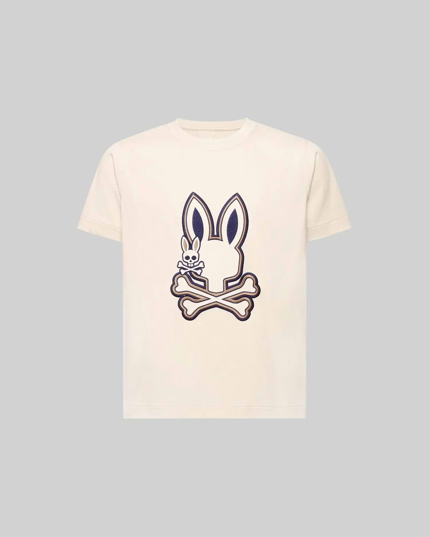 The KIDS WADE GRAPHIC TEE - KT0200054 by Psycho Bunny is a cream-colored 100% cotton tee with a large bunny head and crossbones graphic, plus a small smiling embroidered Bunny on the chest. The background is light gray.