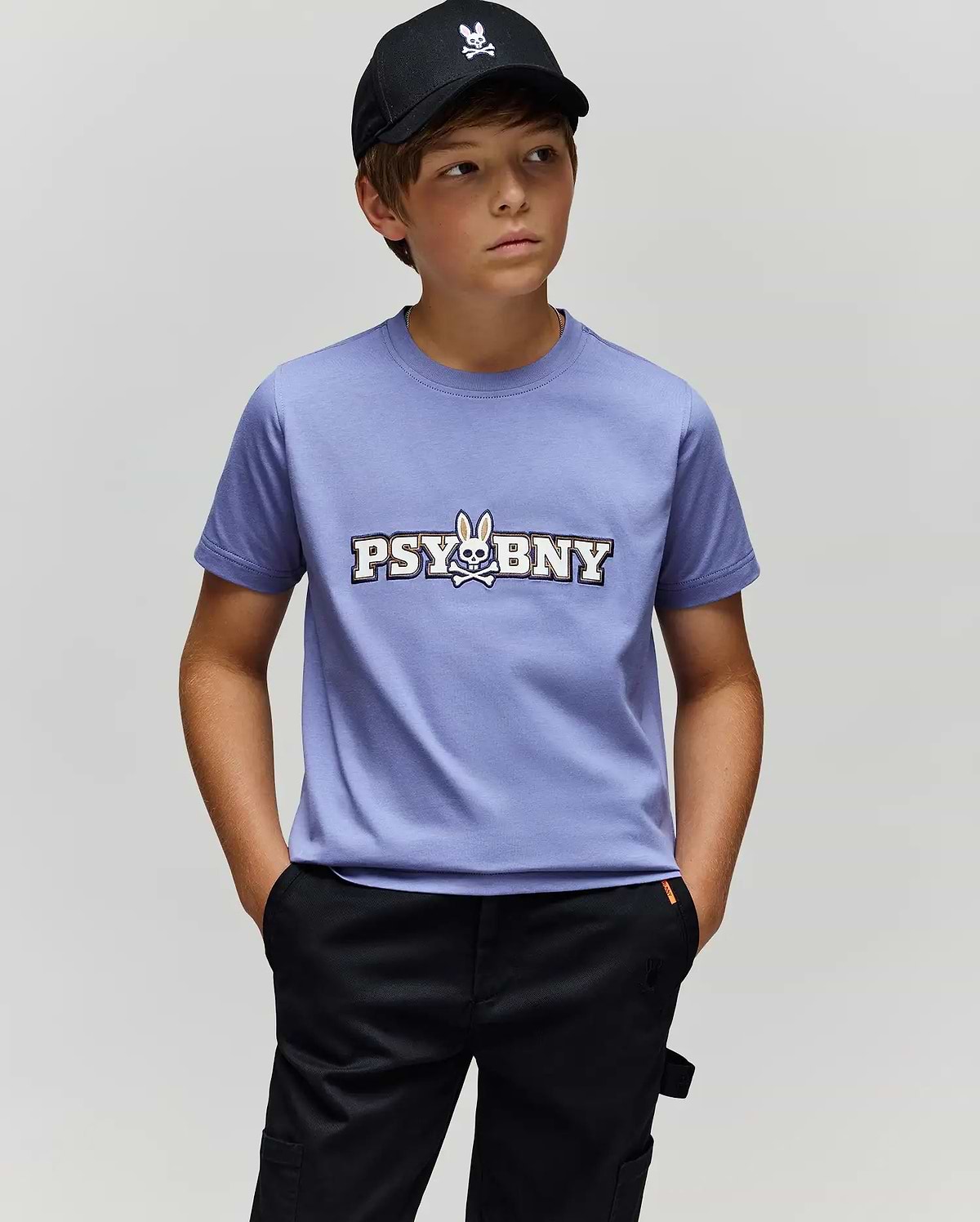 A boy in a black cap, black pants, and the Psycho Bunny KIDS WADE GRAPHIC TEE (KT0200055)—a lavender 100% cotton shirt featuring "PSYBUNY" and a bunny skull logo—stands with hands in pockets against a plain light gray background.