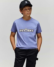 A boy in a black cap, black pants, and the Psycho Bunny KIDS WADE GRAPHIC TEE (KT0200055)—a lavender 100% cotton shirt featuring "PSYBUNY" and a bunny skull logo—stands with hands in pockets against a plain light gray background.