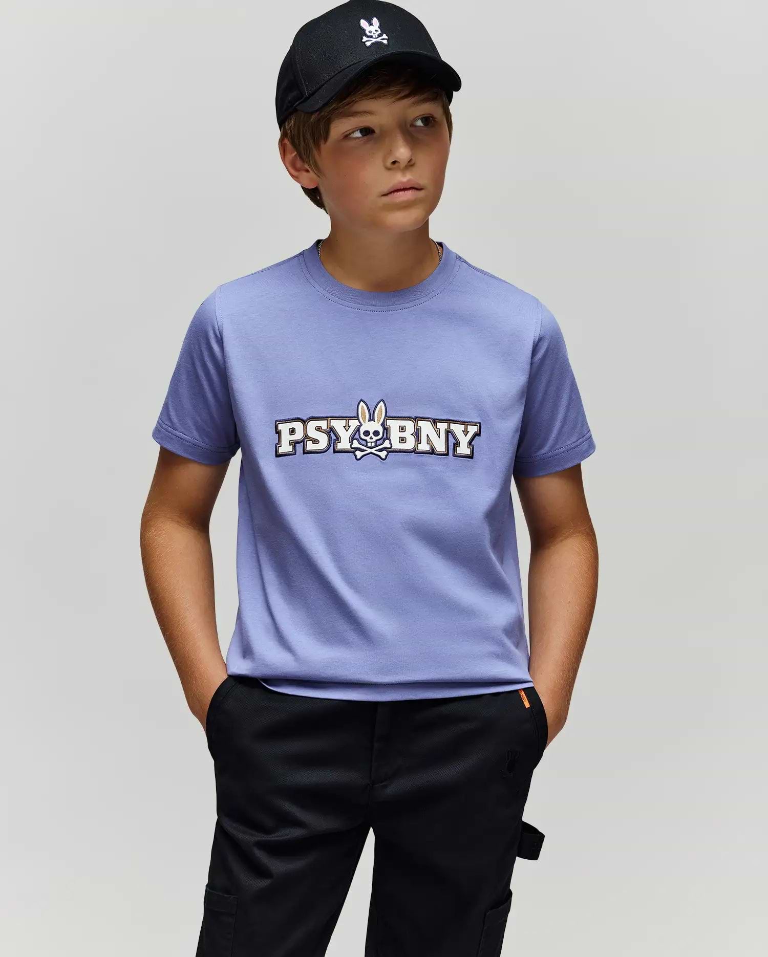 A boy in a black cap, black pants, and the Psycho Bunny KIDS WADE GRAPHIC TEE (KT0200055)—a lavender 100% cotton shirt featuring "PSYBUNY" and a bunny skull logo—stands with hands in pockets against a plain light gray background.