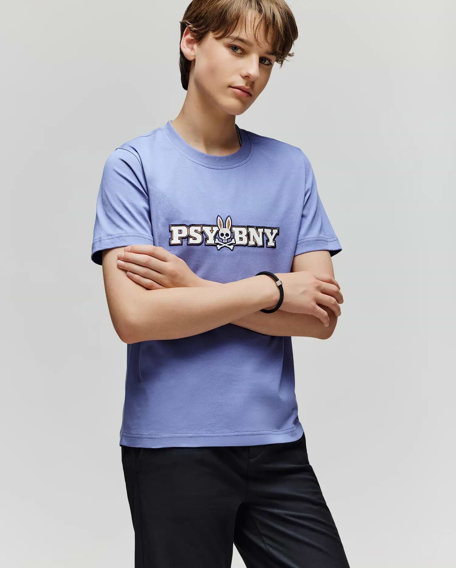 A person with short brown hair stands with arms crossed, wearing the 100% cotton Psycho Bunny KIDS WADE GRAPHIC TEE (KT0200055) in light purple, featuring "PSYBNY" and a bunny graphic, paired with black pants against a plain background.