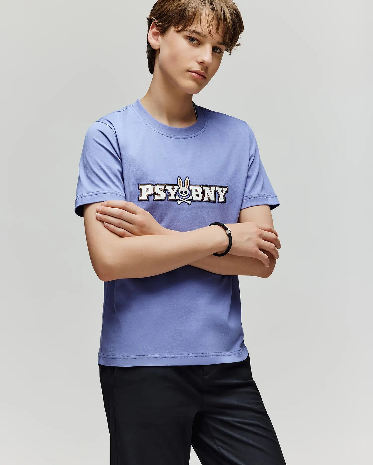 A person with short brown hair stands with arms crossed, wearing the 100% cotton Psycho Bunny KIDS WADE GRAPHIC TEE (KT0200055) in light purple, featuring "PSYBNY" and a bunny graphic, paired with black pants against a plain background.