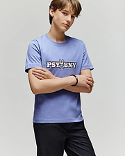 A person with short brown hair stands with arms crossed, wearing the 100% cotton Psycho Bunny KIDS WADE GRAPHIC TEE (KT0200055) in light purple, featuring "PSYBNY" and a bunny graphic, paired with black pants against a plain background.