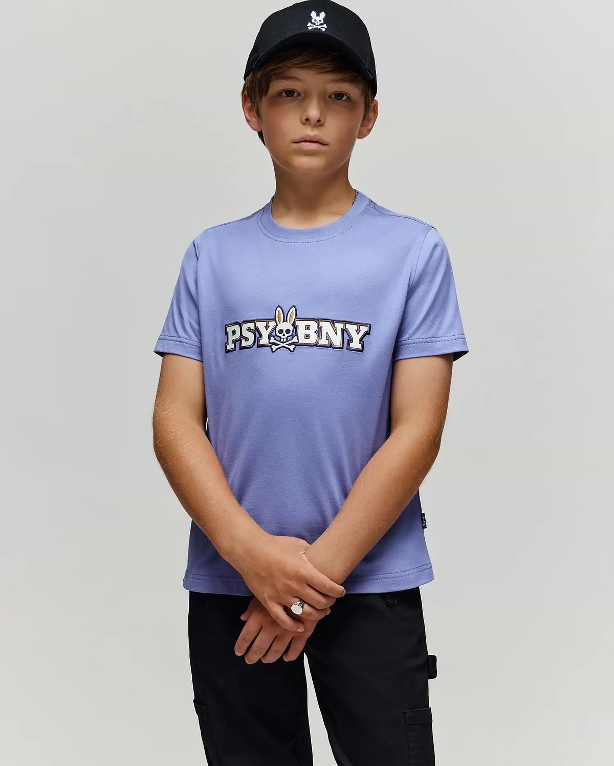 A young boy in a black cap and Psycho Bunny KIDS WADE GRAPHIC TEE (KT0200055) stands against a light gray background, looking at the camera with hands clasped. The lavender tee features 100% cotton, regular fit, cartoon bunny, and "PSYBNY" text.