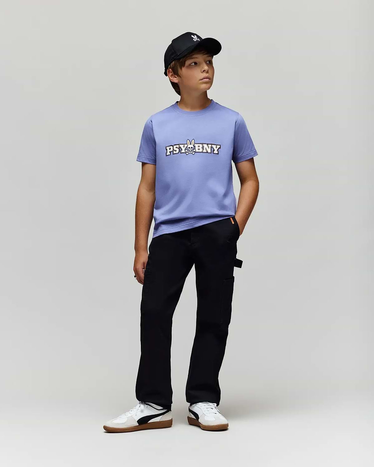 A boy in a black baseball cap, Psycho Bunny KIDS WADE GRAPHIC TEE - KT0200055 (light purple with "PSYBNY" and cartoon bunny), regular fit black pants, and white sneakers stands against a light gray background.