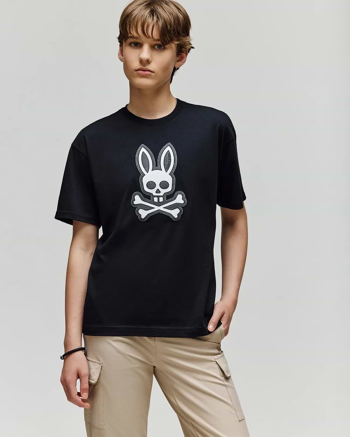 A young person wears the Psycho Bunny KIDS RYDER OVERSIZED FIT TEE (KT0200056), featuring an embroidered Bunny skull and crossbones graphic, with one hand in their beige cargo pants pocket, standing against a plain light background.