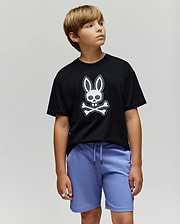 A boy with light brown hair wears the Psycho Bunny KIDS RYDER OVERSIZED FIT TEE (KT0200056) featuring an embroidered Bunny skull and crossbones, paired with light purple shorts. He stands against a plain light background, looking left.
