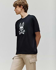 A person with short brown hair wears the Psycho Bunny KIDS RYDER OVERSIZED FIT TEE (KT0200056), featuring an embroidered Bunny skull and crossbones logo, paired with beige cargo pants, standing against a plain light background.
