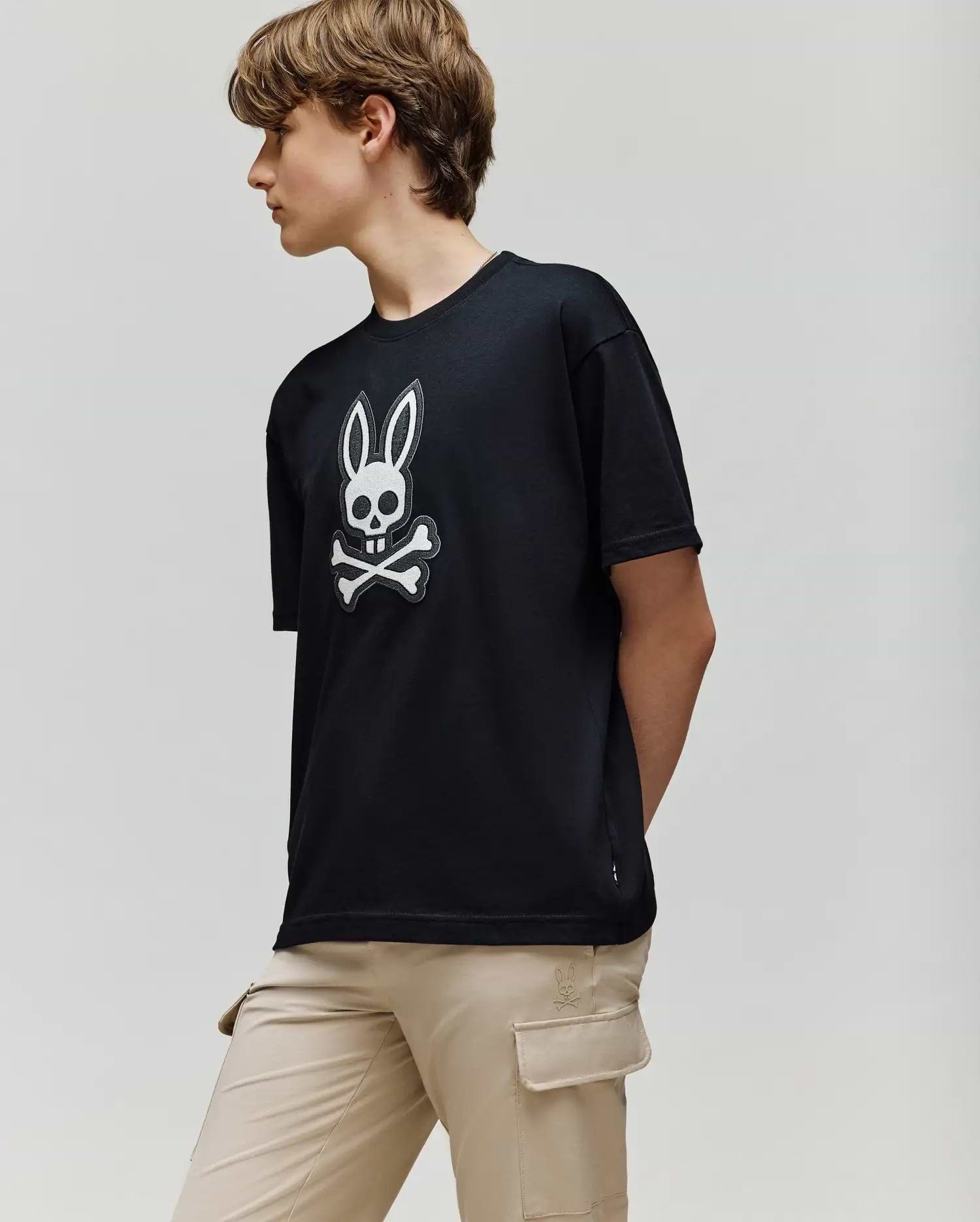 A person with short brown hair wears the Psycho Bunny KIDS RYDER OVERSIZED FIT TEE (KT0200056), featuring an embroidered Bunny skull and crossbones logo, paired with beige cargo pants, standing against a plain light background.