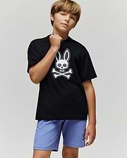 A boy with light brown hair stands confidently in the Psycho Bunny KIDS RYDER OVERSIZED FIT TEE (KT0200056), featuring an embroidered Bunny-skull logo, paired with light purple shorts against a plain gray background.