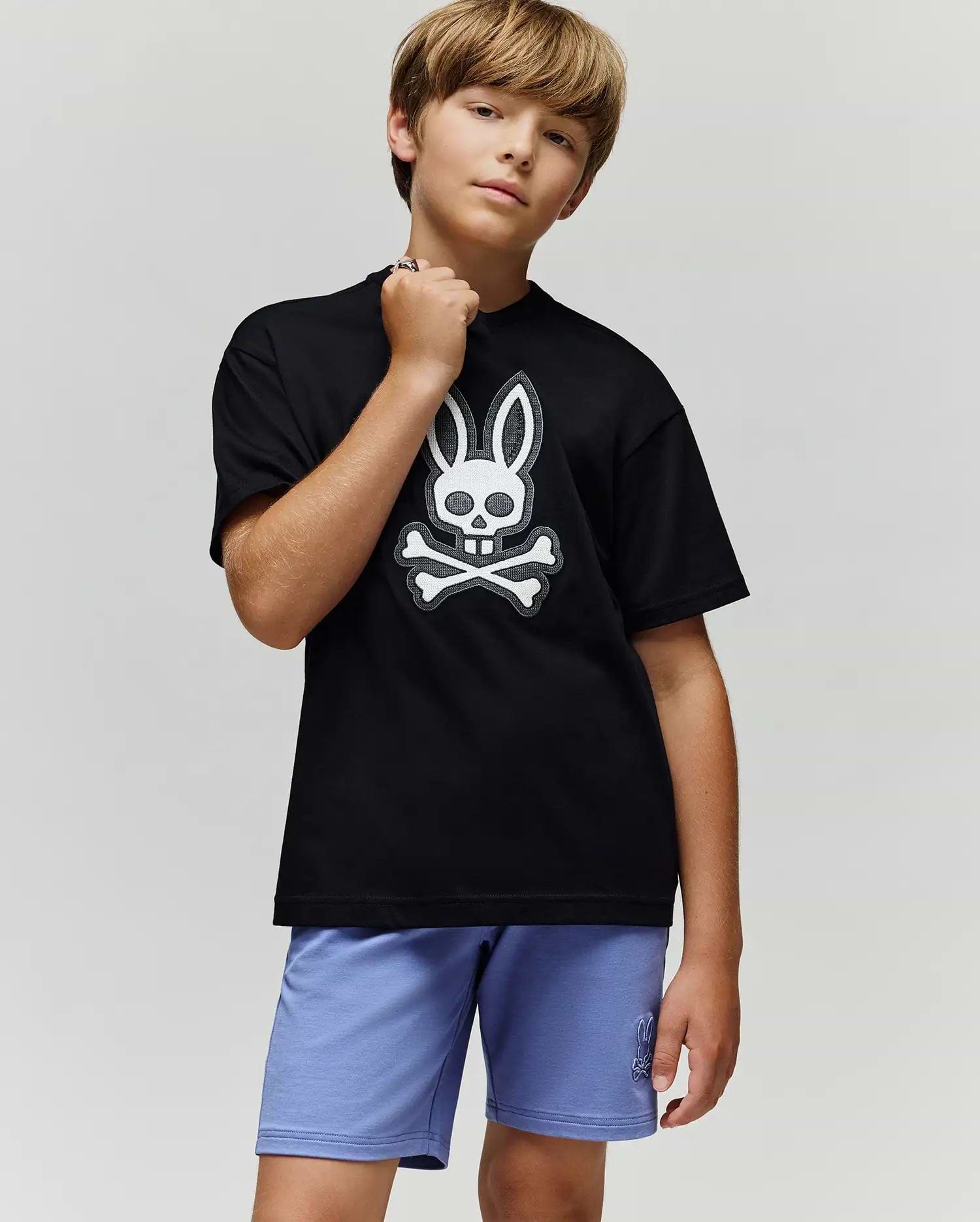 A boy with light brown hair stands confidently in the Psycho Bunny KIDS RYDER OVERSIZED FIT TEE (KT0200056), featuring an embroidered Bunny-skull logo, paired with light purple shorts against a plain gray background.