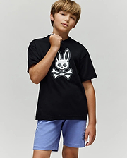 A boy with light brown hair stands confidently in the Psycho Bunny KIDS RYDER OVERSIZED FIT TEE (KT0200056), featuring an embroidered Bunny-skull logo, paired with light purple shorts against a plain gray background.