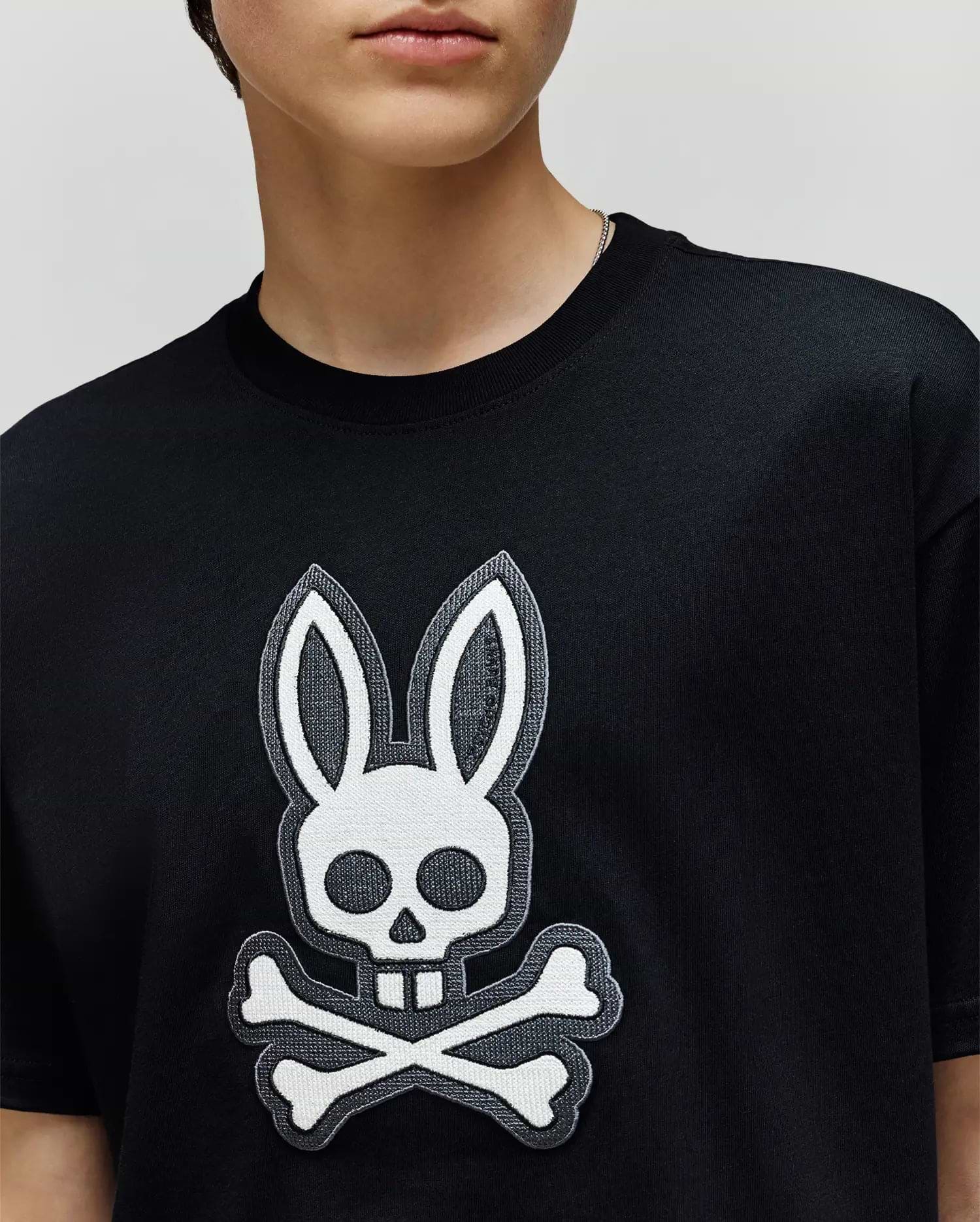 A person wearing the Psycho Bunny KIDS RYDER OVERSIZED FIT TEE (KT0200056), a black premium tee with soft Pima cotton and a large white rabbit head with crossbones graphic. Only the lower half of their face is visible.