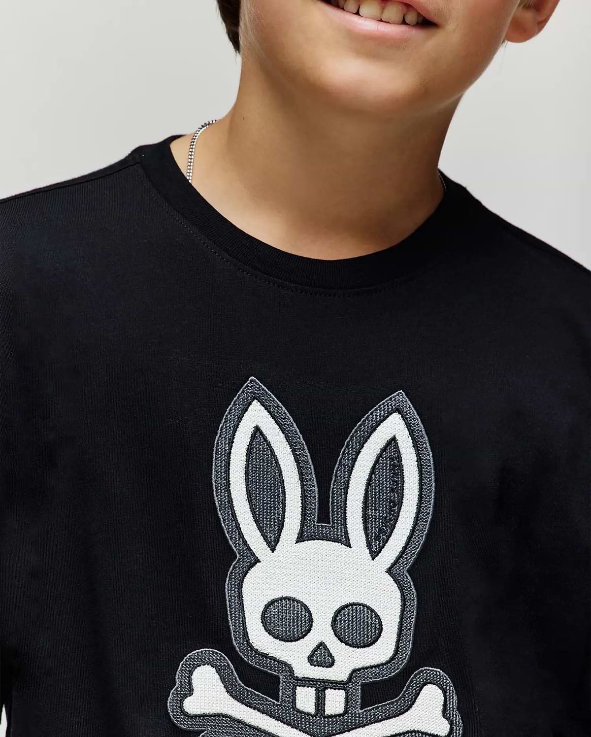A child wears the Psycho Bunny KIDS RYDER OVERSIZED FIT TEE (KT0200056) in black, featuring a bold white skull and crossbones with bunny ears and teeth. Made of soft cotton, it highlights their lower face and upper torso.