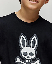 A child wears the Psycho Bunny KIDS RYDER OVERSIZED FIT TEE (KT0200056) in black, featuring a bold white skull and crossbones with bunny ears and teeth. Made of soft cotton, it highlights their lower face and upper torso.