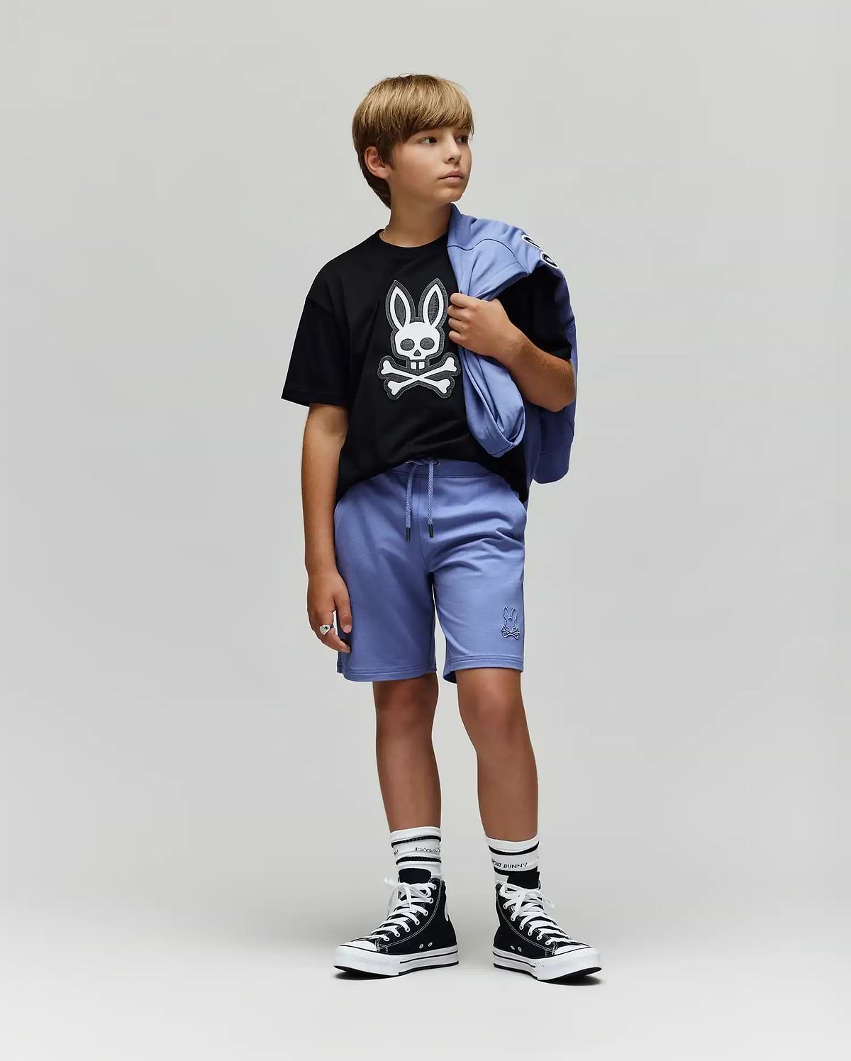 A boy stands against a plain background wearing the Psycho Bunny KIDS RYDER OVERSIZED FIT TEE (KT0200056), featuring an embroidered Bunny and crossbones, paired with light purple shorts, striped socks, black high-top sneakers, and a matching jacket.