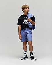 A boy stands against a plain background wearing the Psycho Bunny KIDS RYDER OVERSIZED FIT TEE (KT0200056), featuring an embroidered Bunny and crossbones, paired with light purple shorts, striped socks, black high-top sneakers, and a matching jacket.