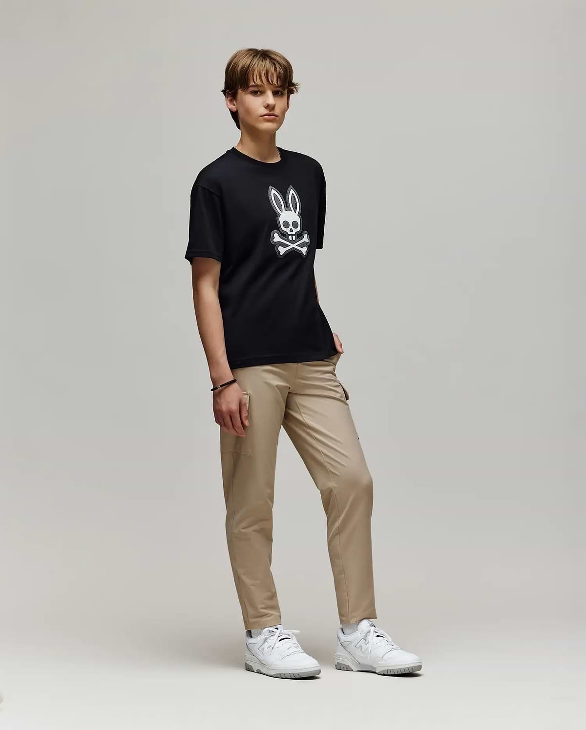 A young person with short hair stands against a plain background, wearing the Psycho Bunny KIDS RYDER OVERSIZED FIT TEE (KT0200056) featuring a white cartoon skull and bunny ears graphic, paired with beige pants and white sneakers.