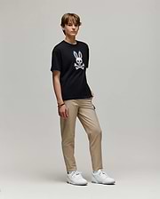 A young person with short hair stands against a plain background, wearing the Psycho Bunny KIDS RYDER OVERSIZED FIT TEE (KT0200056) featuring a white cartoon skull and bunny ears graphic, paired with beige pants and white sneakers.