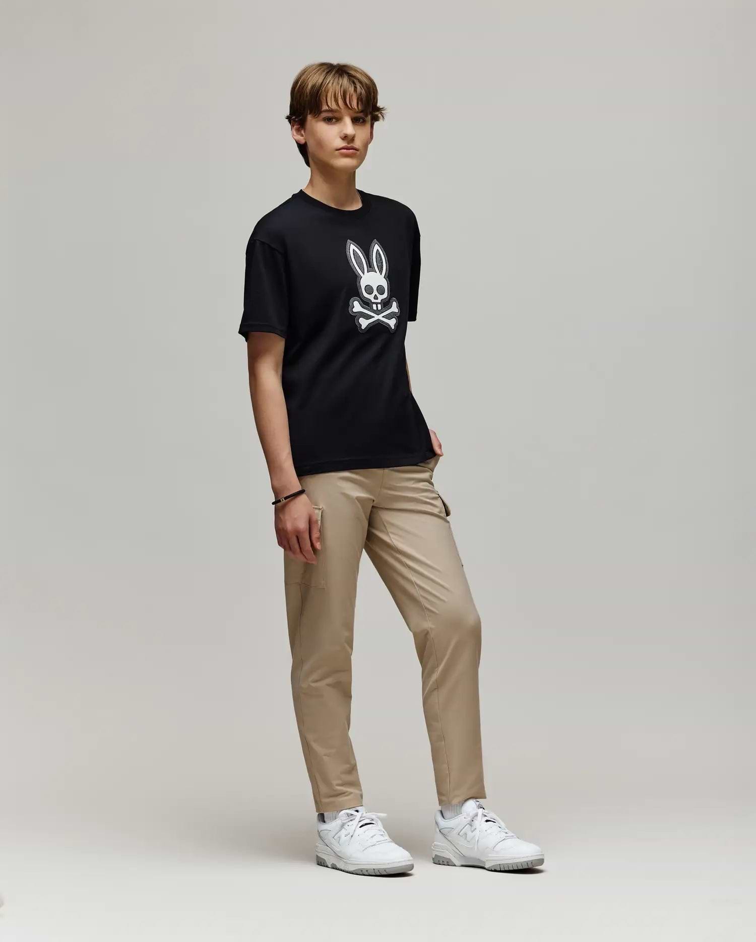 A young person with short hair stands against a plain background, wearing the Psycho Bunny KIDS RYDER OVERSIZED FIT TEE (KT0200056) featuring a white cartoon skull and bunny ears graphic, paired with beige pants and white sneakers.