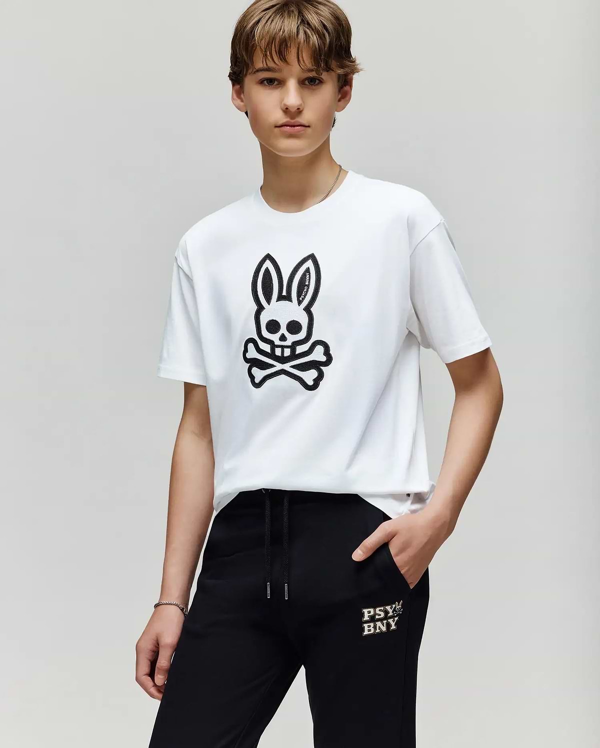 A person with short brown hair wears the Psycho Bunny KIDS RYDER OVERSIZED FIT TEE (KT0200056) with a black embroidered Bunny skull graphic, paired with black "PSY BNXY" pants, standing against a plain light background.