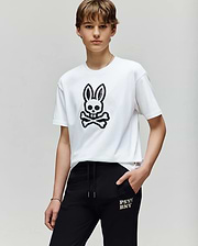 A person with short brown hair wears the Psycho Bunny KIDS RYDER OVERSIZED FIT TEE (KT0200056) with a black embroidered Bunny skull graphic, paired with black "PSY BNXY" pants, standing against a plain light background.
