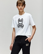 A person with short brown hair wears the Psycho Bunny KIDS RYDER OVERSIZED FIT TEE (KT0200056) with a black embroidered Bunny skull graphic, paired with black "PSY BNXY" pants, standing against a plain light background.