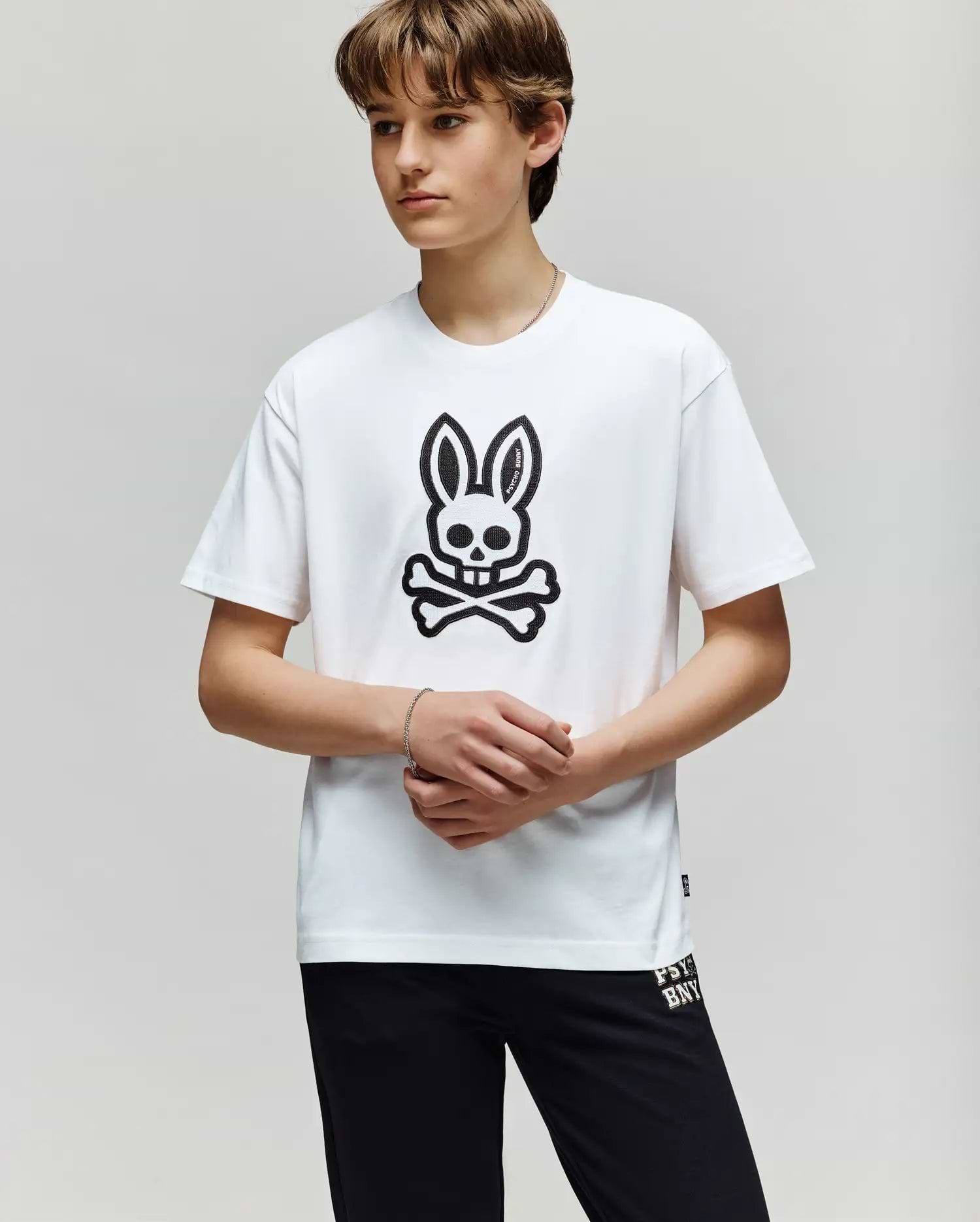 A young person stands against a plain background, wearing the Psycho Bunny KIDS RYDER OVERSIZED FIT TEE - KT0200056 with a large black bunny skull and crossbones graphic, paired with black pants. Their hands are loosely clasped in front.