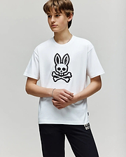 A young person stands against a plain background, wearing the Psycho Bunny KIDS RYDER OVERSIZED FIT TEE - KT0200056 with a large black bunny skull and crossbones graphic, paired with black pants. Their hands are loosely clasped in front.