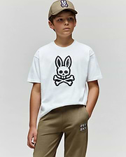 A boy stands against a plain background, wearing a khaki cap, khaki pants, and the Psycho Bunny KIDS RYDER OVERSIZED FIT TEE (KT0200056), featuring premium Pima cotton and an embroidered Bunny skull and crossbones graphic.