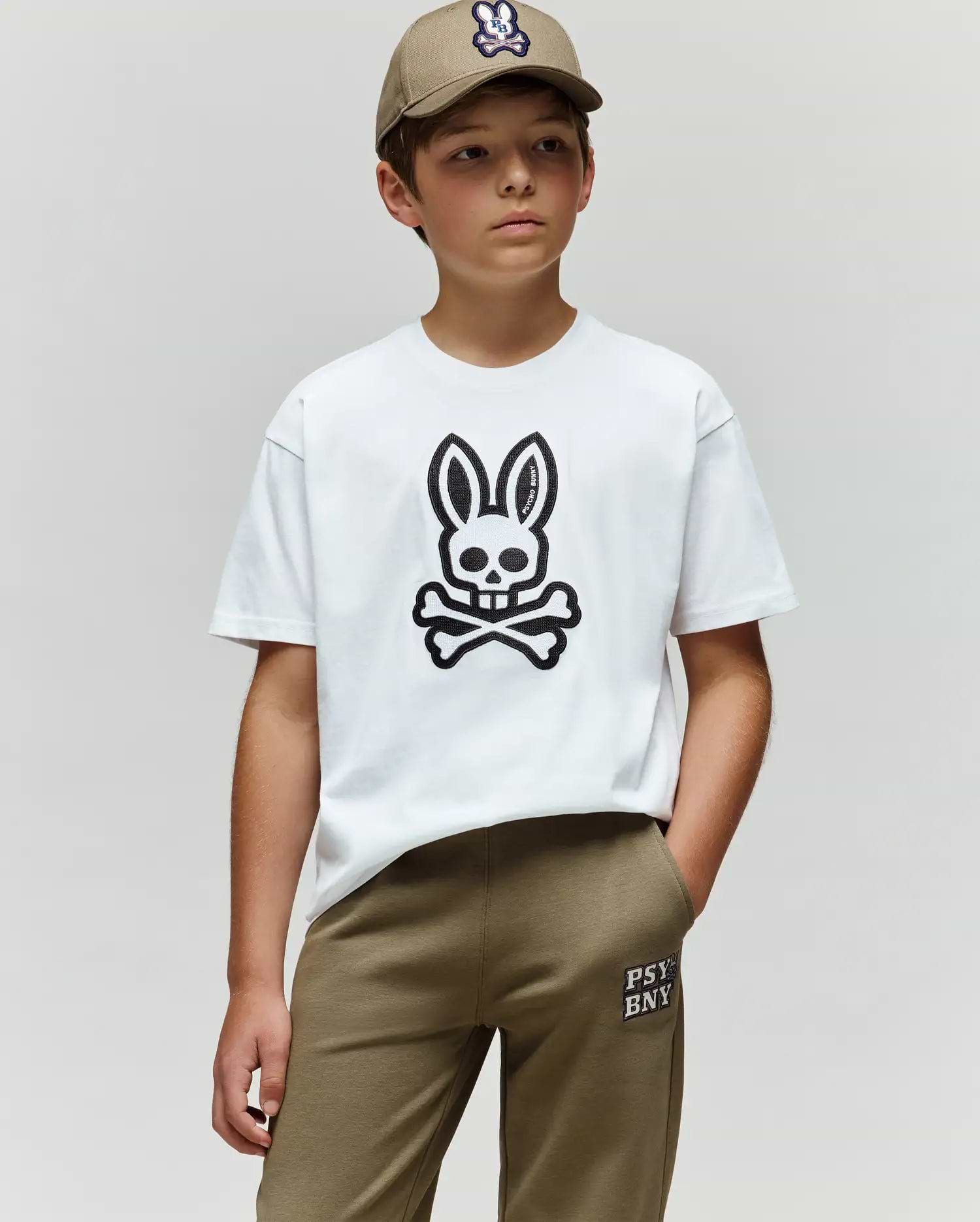 A boy stands against a plain background, wearing a khaki cap, khaki pants, and the Psycho Bunny KIDS RYDER OVERSIZED FIT TEE (KT0200056), featuring premium Pima cotton and an embroidered Bunny skull and crossbones graphic.