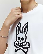 A person wears the kids' Ryder oversized fit tee (KT0200056) by Psycho Bunny, featuring a white base with a black skull and crossbones bunny design; their hand near their neck reveals a silver chain bracelet.
