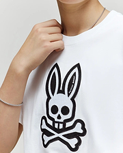 A person wears the kids' Ryder oversized fit tee (KT0200056) by Psycho Bunny, featuring a white base with a black skull and crossbones bunny design; their hand near their neck reveals a silver chain bracelet.