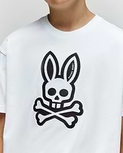 A person wearing the Psycho Bunny KIDS RYDER OVERSIZED FIT TEE (KT0200056), made of soft Pima cotton, featuring a large black cartoon bunny head with skull-like eyes and nose above crossed bones on the front.
