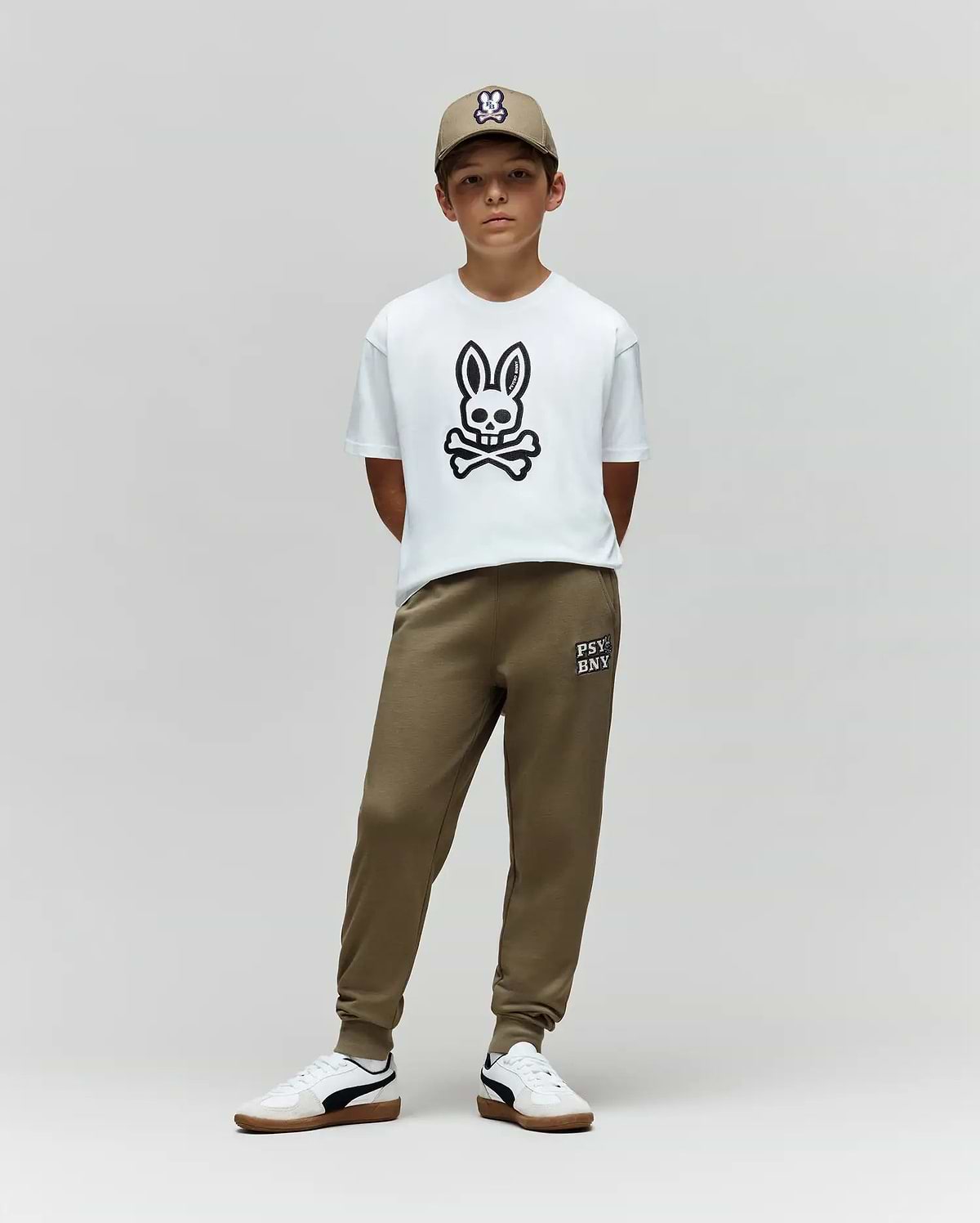 A young boy stands against a plain background, wearing the Psycho Bunny KIDS RYDER OVERSIZED FIT TEE - KT0200056 with a large bunny and crossbones graphic, olive green joggers, white sneakers, and an olive green cap with an embroidered Bunny logo.