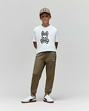 A young boy stands against a plain background, wearing the Psycho Bunny KIDS RYDER OVERSIZED FIT TEE - KT0200056 with a large bunny and crossbones graphic, olive green joggers, white sneakers, and an olive green cap with an embroidered Bunny logo.
