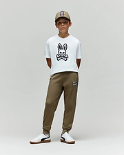 A young boy stands against a plain background, wearing the Psycho Bunny KIDS RYDER OVERSIZED FIT TEE - KT0200056 with a large bunny and crossbones graphic, olive green joggers, white sneakers, and an olive green cap with an embroidered Bunny logo.
