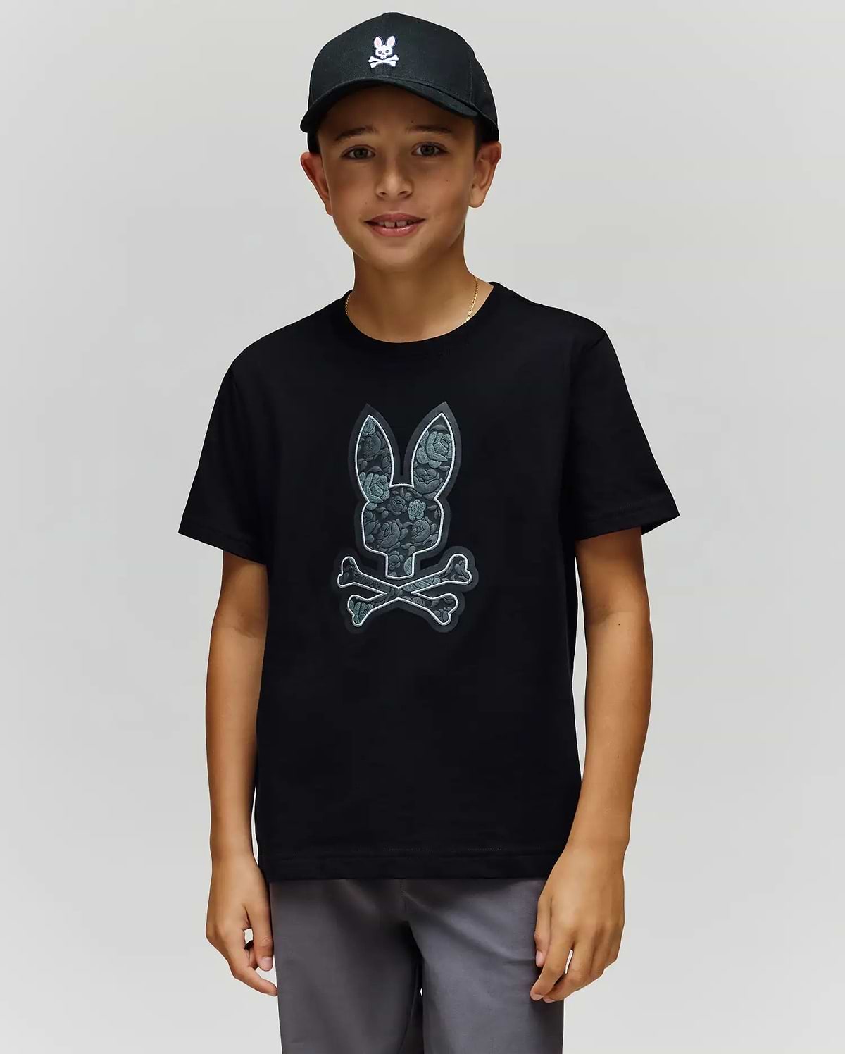 A boy in a black cap and the Psycho Bunny KIDS ROWEN BUNNY GRAPHIC TEE (KT0200058), crafted from soft Pima cotton in Peru, stands facing the camera against a plain light background. (1)