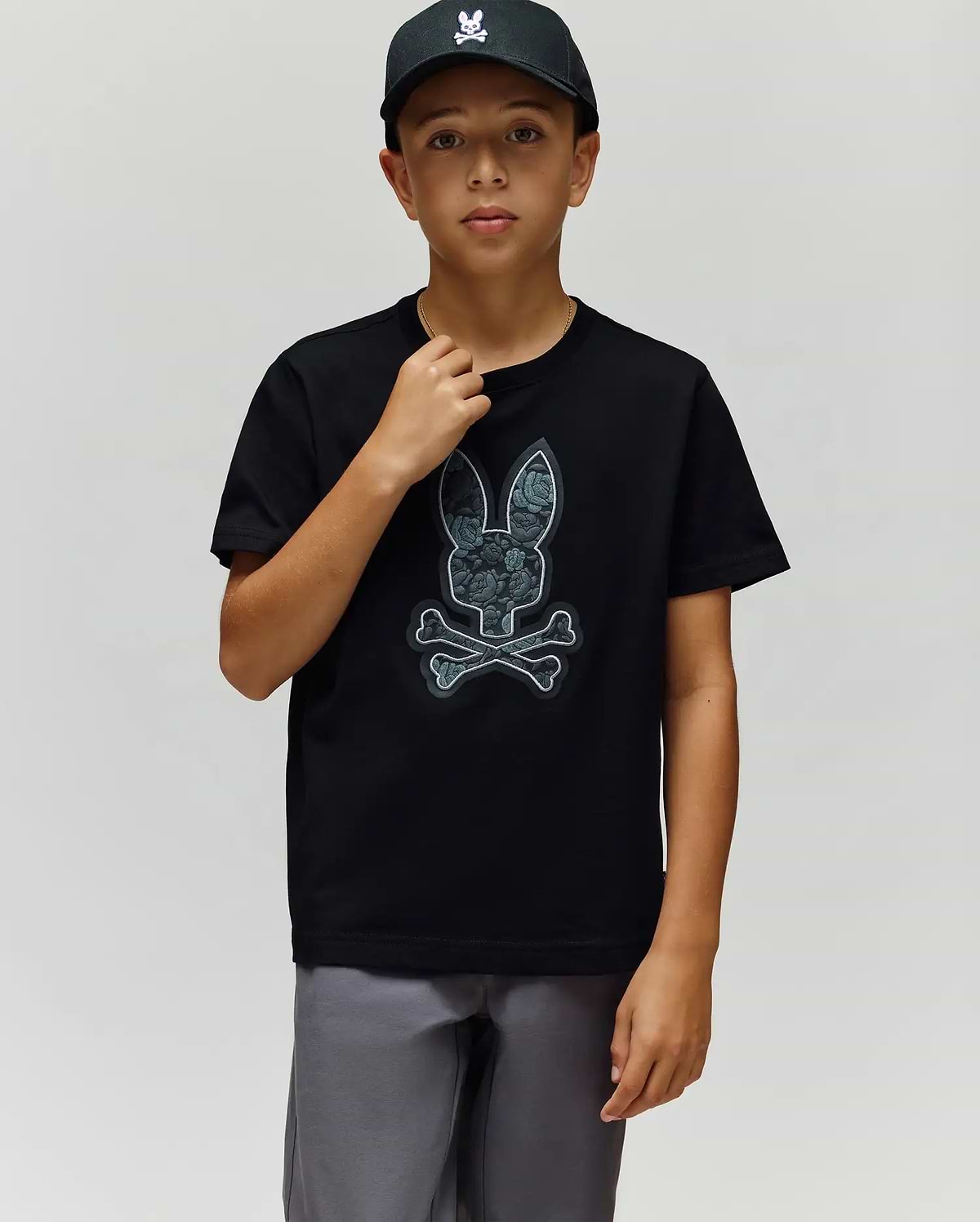 A boy in a black cap, gray shorts, and the Psycho Bunny KIDS ROWEN BUNNY GRAPHIC TEE (KT0200058), made from soft Pima cotton in Peru, stands against a plain light background. (3)