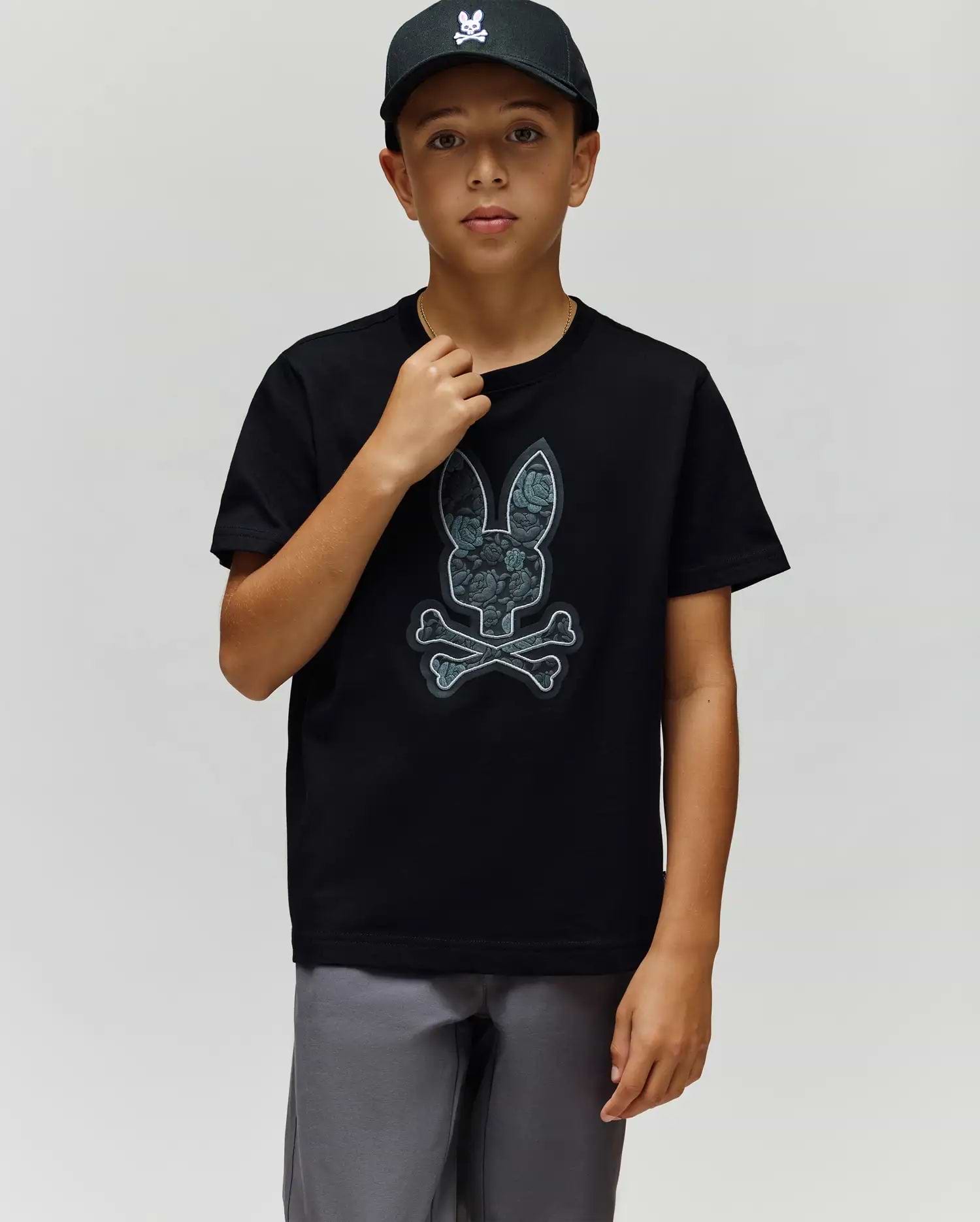 A boy in a black cap, gray shorts, and the Psycho Bunny KIDS ROWEN BUNNY GRAPHIC TEE (KT0200058), made from soft Pima cotton in Peru, stands against a plain light background. (3)