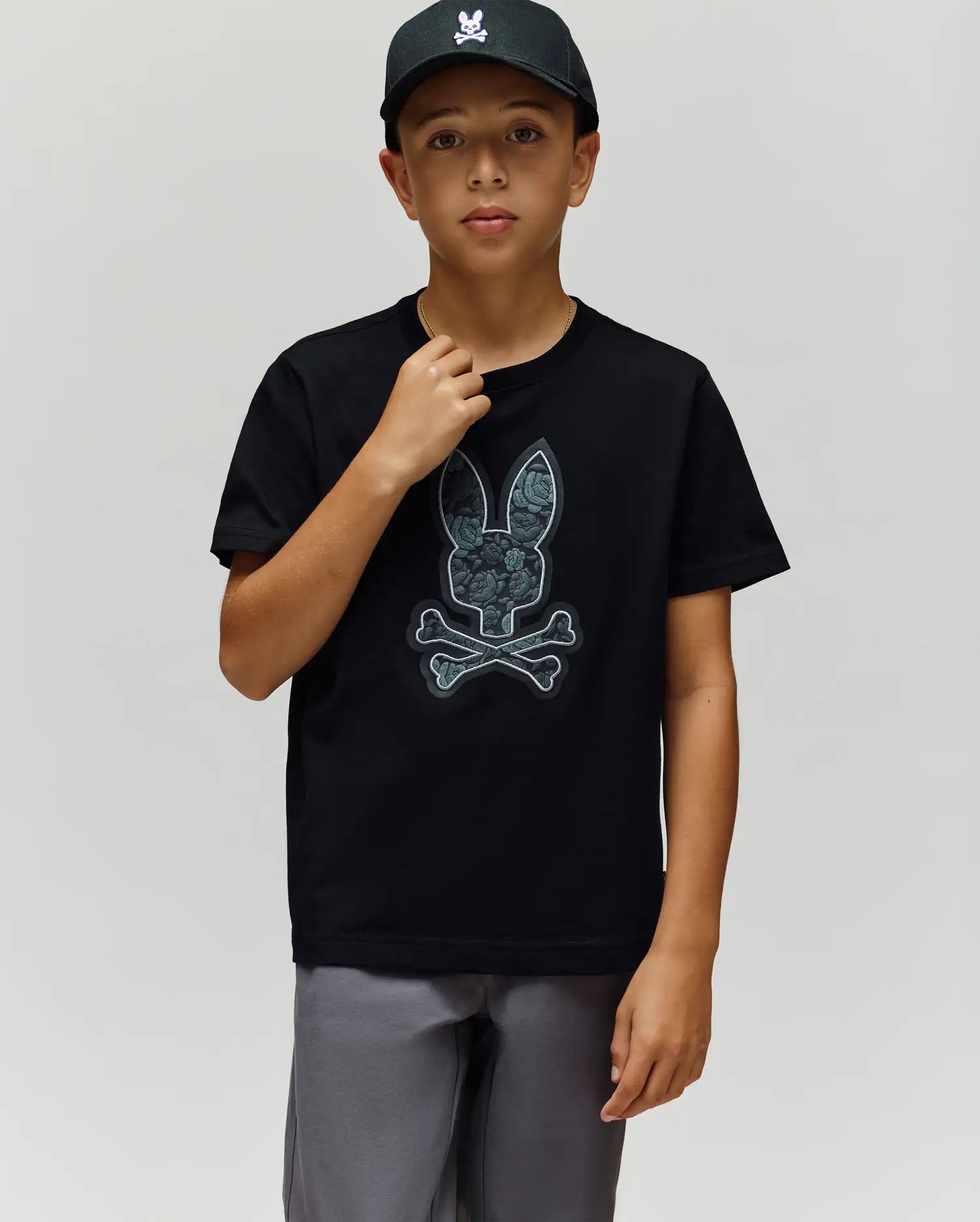 A boy in a black cap, gray shorts, and the Psycho Bunny KIDS ROWEN BUNNY GRAPHIC TEE (KT0200058), made from soft Pima cotton in Peru, stands against a plain light background. (3)